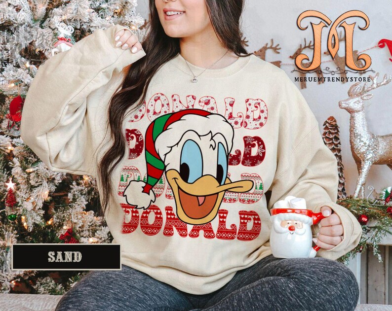 May include: Sand-colored sweatshirt featuring a graphic of Donald Duck in a Santa hat with the name "DONALD" in a festive font. The sweatshirt is part of a Christmas-themed collection.