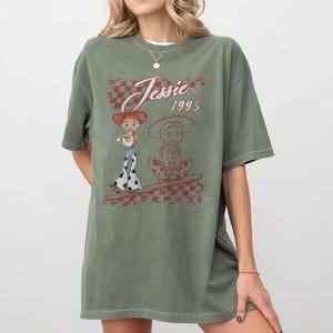 K&ouml;nnte beinhalten: Olivgr&uuml;nes T-Shirt mit einer Grafik von Jessie aus Toy Story. Das Design enth&auml;lt den Text "Jessie 1995" in einem rot-wei&szlig; karierten Muster. Das Shirt hat kurze &Auml;rmel und eine lockere Passform.