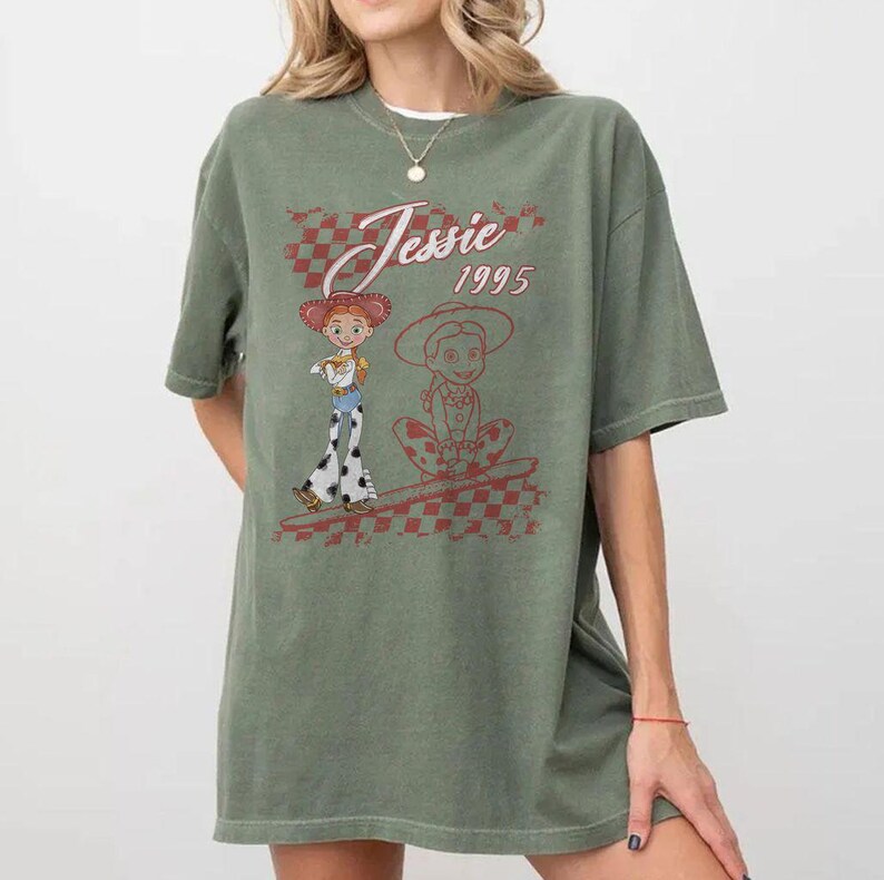 K&ouml;nnte beinhalten: Olivgr&uuml;nes T-Shirt mit einer Grafik von Jessie aus Toy Story. Das Design enth&auml;lt den Text "Jessie 1995" in einem rot-wei&szlig; karierten Muster. Das Shirt hat kurze &Auml;rmel und eine lockere Passform.