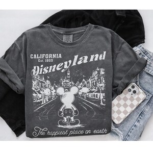 以下が含まれることがあります： グレーの長袖Tシャツ。かすれた風合いのディズニーランドのグラフィック。ミッキーマウス、テキスト「CALIFORNIA Disneyland Est. 1955」、および「The happiest place on earth」が特徴です。市松模様のケースの電話とデニムジーンズも表示されています。