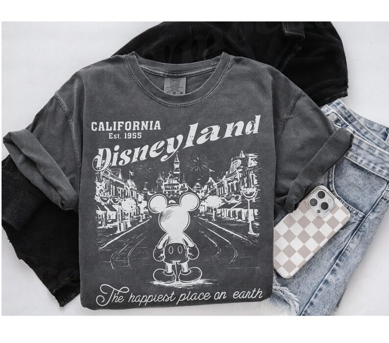 以下が含まれることがあります： グレーの長袖Tシャツ。かすれた風合いのディズニーランドのグラフィック。ミッキーマウス、テキスト「CALIFORNIA Disneyland Est. 1955」、および「The happiest place on earth」が特徴です。市松模様のケースの電話とデニムジーンズも表示されています。