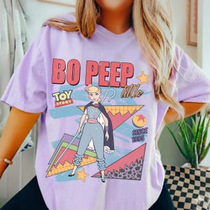 May include: A light purple t-shirt featuring a graphic of Bo Peep from Toy Story. The design includes the text "BO PEEP" in red, "TOY STORY" in blue, and "Little" in white. The shirt also has a cartoon image of Bo Peep.