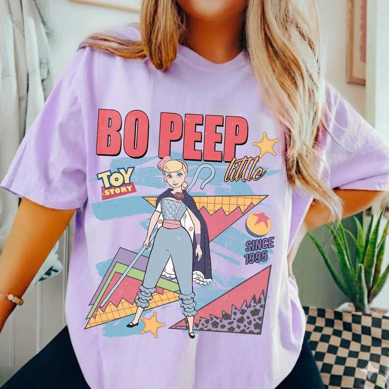 May include: A light purple t-shirt featuring a graphic of Bo Peep from Toy Story. The design includes the text "BO PEEP" in red, "TOY STORY" in blue, and "Little" in white. The shirt also has a cartoon image of Bo Peep.