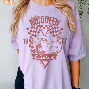 May include: A purple t-shirt with a brown graphic of a race car with the text "Lightning McQueen" and "Cars" on it. The car is number 95 and the text "19" and "77" are on either side of the car.