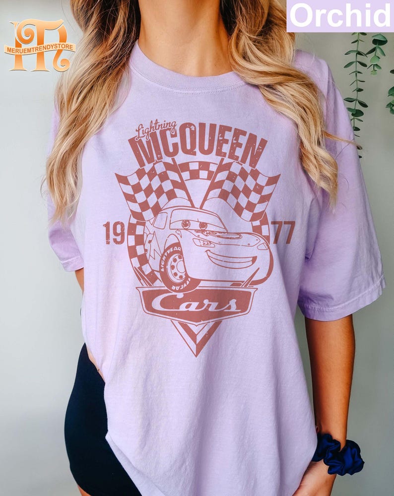 May include: A purple t-shirt with a brown graphic of a race car with the text "Lightning McQueen" and "Cars" on it. The car is number 95 and the text "19" and "77" are on either side of the car.