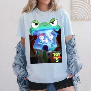 May include: Pale blue t-shirt with a green frog-shaped frame displaying a scene from "Toy Story 5". The frame shows Woody, Buzz Lightyear, and Jessie. The shirt is made of a soft material.