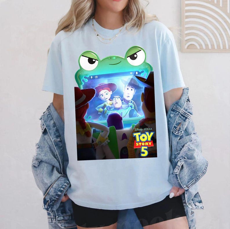 May include: Pale blue t-shirt with a green frog-shaped frame displaying a scene from "Toy Story 5". The frame shows Woody, Buzz Lightyear, and Jessie. The shirt is made of a soft material.