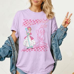 K&ouml;nnte beinhalten: Ein lila T-Shirt mit einer Grafik von Bo Peep aus dem Animationsfilm von 1995. Das Design zeigt zwei Bo Peep-Illustrationen, ein rosa-wei&szlig;es Schachmuster und den Text "Bo Peep 1995" in einer Retro-Schriftart.