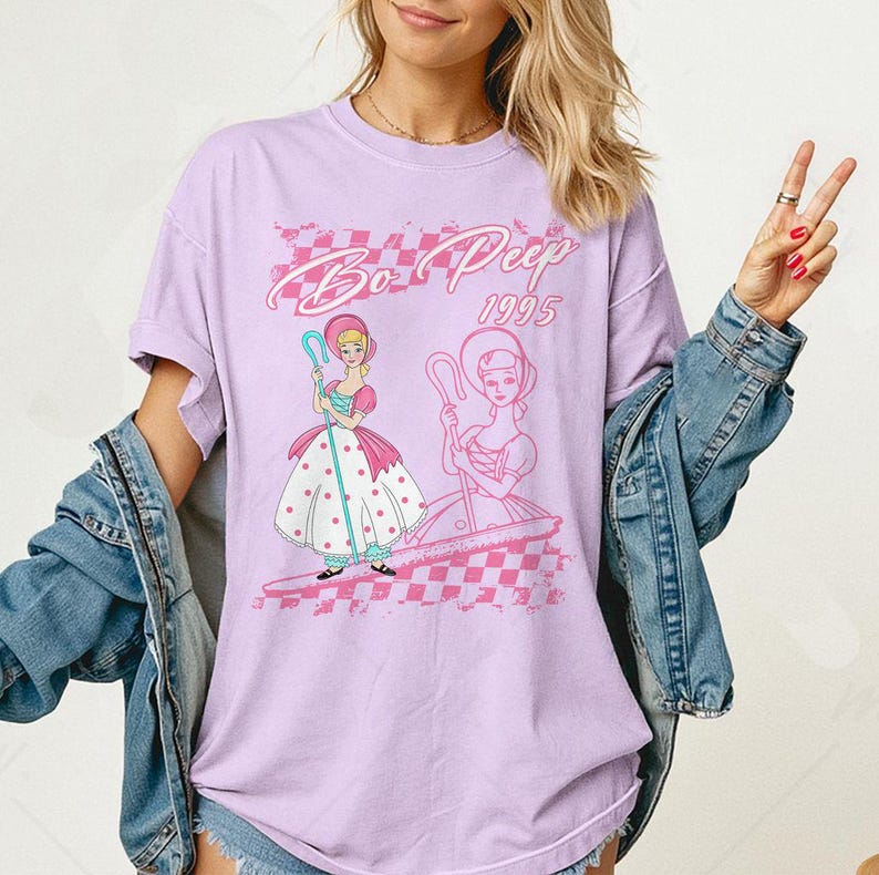 K&ouml;nnte beinhalten: Ein lila T-Shirt mit einer Grafik von Bo Peep aus dem Animationsfilm von 1995. Das Design zeigt zwei Bo Peep-Illustrationen, ein rosa-wei&szlig;es Schachmuster und den Text "Bo Peep 1995" in einer Retro-Schriftart.