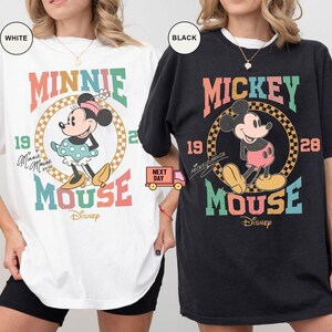 May include: Two oversized t-shirts featuring vintage-style graphics of Mickey and Minnie Mouse. The Mickey Mouse shirt is black with a colorful graphic that says "Mickey Mouse" and "19 28". The Minnie Mouse shirt is white with a colorful graphic that says "Minnie Mouse" and "19 28".