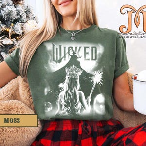 May include: Moss green t-shirt featuring a graphic from the musical "Wicked". The design includes the word "Wicked" above an illustration of a woman with a crown and wand. The shirt is a comfortable fit.