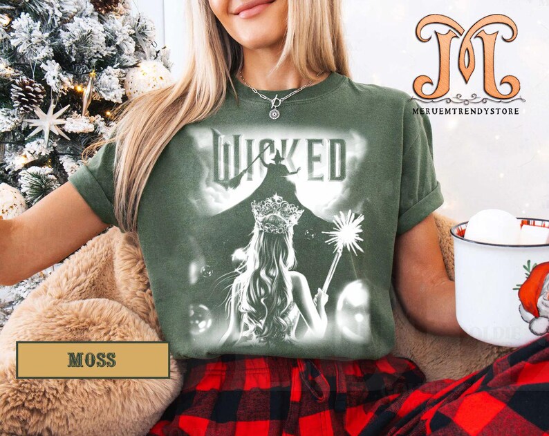 May include: Moss green t-shirt featuring a graphic from the musical "Wicked". The design includes the word "Wicked" above an illustration of a woman with a crown and wand. The shirt is a comfortable fit.