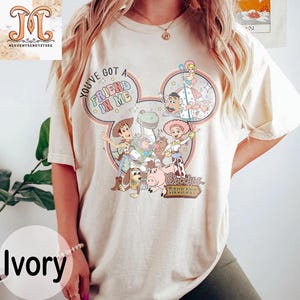 Puede incluir: Una camiseta beige claro con una imagen de dibujos animados de personajes de la pel&iacute;cula Toy Story. La imagen tiene forma de oreja de Mickey Mouse e incluye el texto "You've got a friend in me" y "Woody's Roundup".