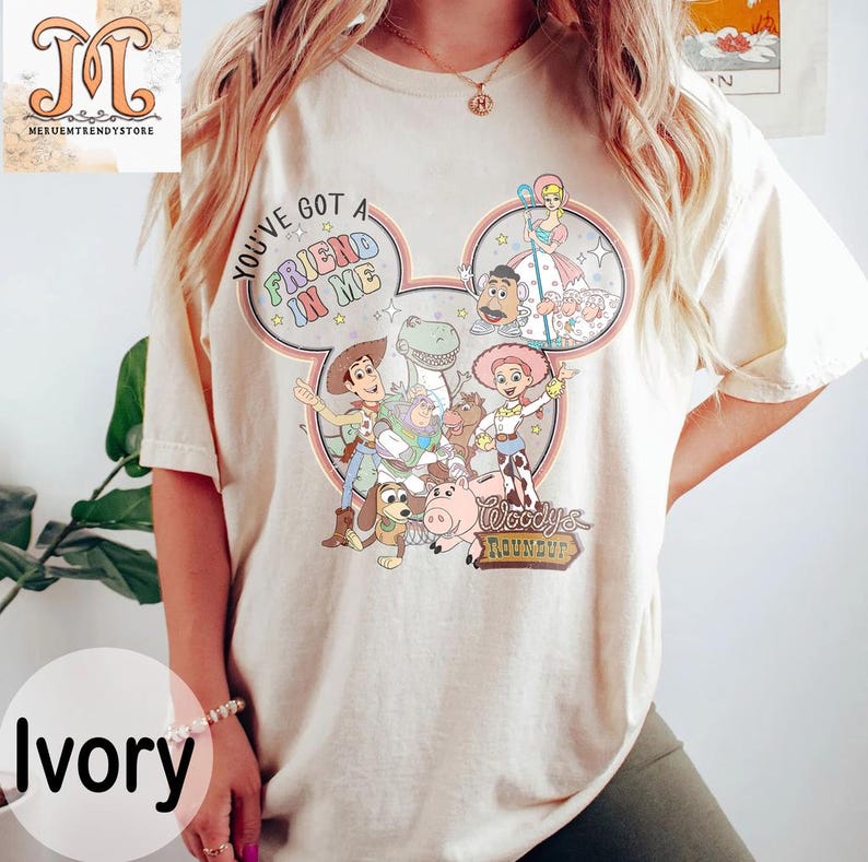 Puede incluir: Una camiseta beige claro con una imagen de dibujos animados de personajes de la pel&iacute;cula Toy Story. La imagen tiene forma de oreja de Mickey Mouse e incluye el texto "You've got a friend in me" y "Woody's Roundup".