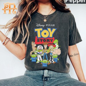 May include: Dark gray t-shirt featuring the Disney Pixar Toy Story logo and characters. The design includes Woody, Buzz Lightyear, and other characters from the animated film. The shirt has a vintage, worn look.