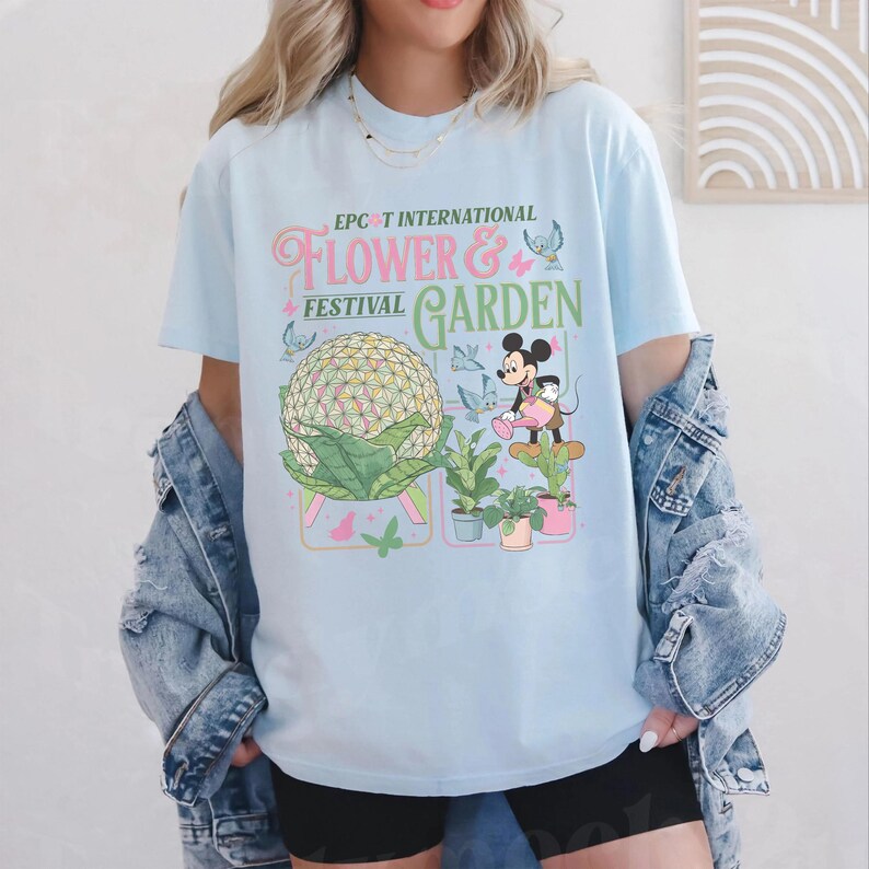 May include: Light blue t-shirt featuring the text "EPCOT INTERNATIONAL FLOWER & GARDEN FESTIVAL" with a graphic of Spaceship Earth and Mickey Mouse. The shirt has a crew neck and short sleeves.