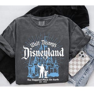 Puede incluir: Una camiseta gris de manga larga con un gr&aacute;fico desgastado del castillo de Disneyland y el texto "Walt Disney's Disneyland". El gr&aacute;fico es en blanco y azul claro. La camiseta tambi&eacute;n tiene el texto "The Happiest Place On Earth EST. 1955".