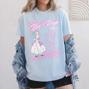 K&ouml;nnte beinhalten: Hellblaues T-Shirt mit einem Bo Peep-Motiv und dem Text "Bo Peep 1995". Das Design beinhaltet ein rosa-wei&szlig;es Schachmuster und eine Cartoon-Illustration von Bo Peep mit einem Hirtenstab. Das T-Shirt hat kurze &Auml;rmel.