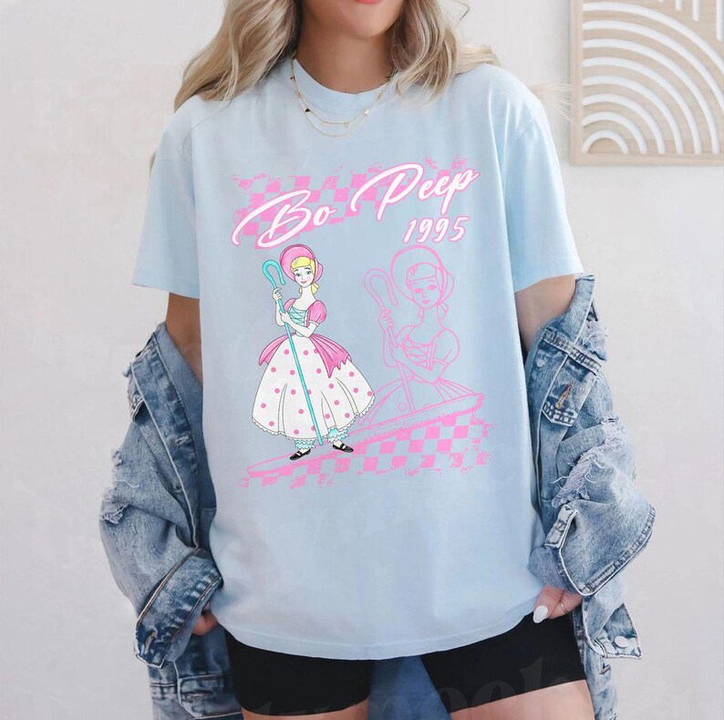 K&ouml;nnte beinhalten: Hellblaues T-Shirt mit einem Bo Peep-Motiv und dem Text "Bo Peep 1995". Das Design beinhaltet ein rosa-wei&szlig;es Schachmuster und eine Cartoon-Illustration von Bo Peep mit einem Hirtenstab. Das T-Shirt hat kurze &Auml;rmel.