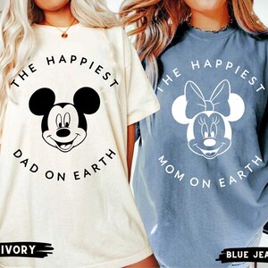 May include: Two t-shirts, one ivory and one blue jean, featuring Mickey and Minnie Mouse. The ivory shirt says "The Happiest Dad on Earth." The blue jean shirt says "The Happiest Mom on Earth."