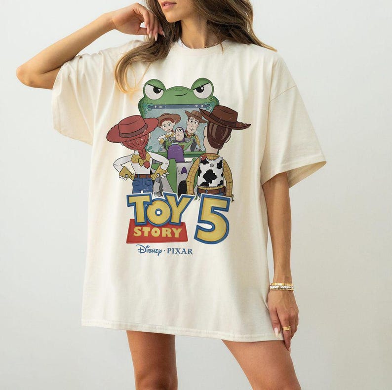 May include: Cream-colored t-shirt featuring a graphic from the movie "Toy Story 5". The design includes cartoon characters, the movie title, and the Disney Pixar logo. The shirt has short sleeves and a relaxed fit.