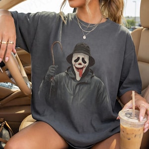 Könnte beinhalten: Dunkelgraues T-Shirt mit einer Grafik einer Figur aus dem Film Scream. Die Figur trägt einen schwarzen Hut und Mantel und hält einen Haken. Die Maske der Figur ist weiß mit einer roten Zunge. Das T-Shirt hat eine lockere Passform.