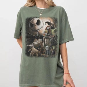 May include: An olive green t-shirt featuring a graphic from "The Nightmare Before Christmas." The design includes Jack Skellington, Sally, Zero, and other characters. The shirt has a crew neck and short sleeves.