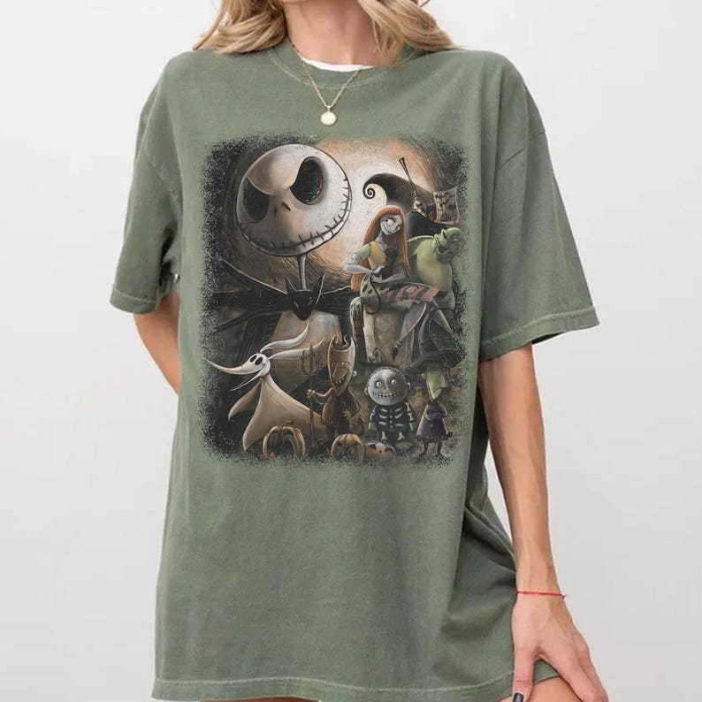 May include: An olive green t-shirt featuring a graphic from "The Nightmare Before Christmas." The design includes Jack Skellington, Sally, Zero, and other characters. The shirt has a crew neck and short sleeves.