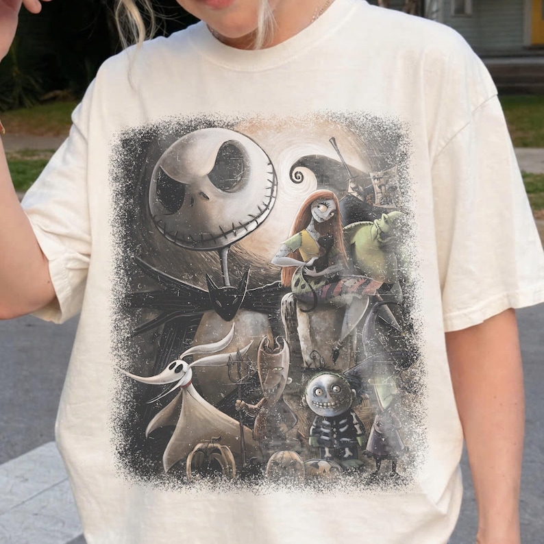May include: Cream-colored t-shirt featuring a graphic print of characters from The Nightmare Before Christmas. The design includes Jack Skellington, Sally, Zero, and other figures in a dark, spooky setting. The shirt has a classic crew neck and short sleeves.