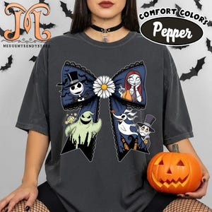 Peut inclure: T-shirt gris fonc&eacute; avec un grand motif de n&oelig;ud orn&eacute; de personnages de L'&Eacute;trange No&euml;l de Monsieur Jack. Le n&oelig;ud a une marguerite blanche au centre et est bord&eacute; de dentelle noire. Le t-shirt porte &eacute;galement les mots "Comfort Colors Pepper".