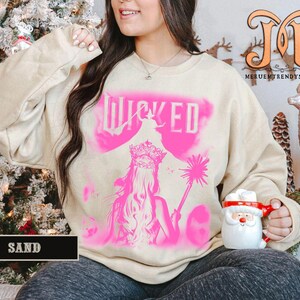 May include: A sand-colored sweatshirt with a pink graphic of a woman with a crown and the word "WICKED". The sweatshirt has long sleeves and a crew neck. The image is a close-up shot.