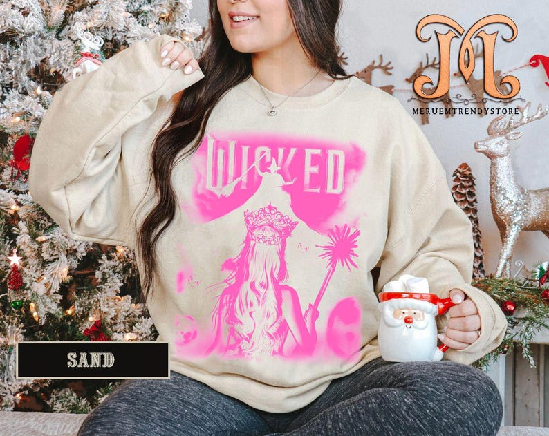 May include: A sand-colored sweatshirt with a pink graphic of a woman with a crown and the word "WICKED". The sweatshirt has long sleeves and a crew neck. The image is a close-up shot.