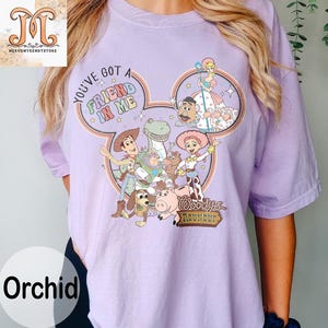 Puede incluir: Una camiseta morada con una imagen de dibujos animados de Woody y sus amigos de Toy Story. La imagen tiene la forma de una oreja de Mickey Mouse e incluye el texto "You've got a friend in me" y "Woody's Roundup".