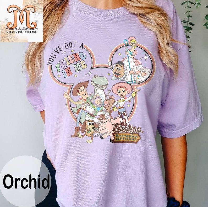 Puede incluir: Una camiseta morada con una imagen de dibujos animados de Woody y sus amigos de Toy Story. La imagen tiene la forma de una oreja de Mickey Mouse e incluye el texto "You've got a friend in me" y "Woody's Roundup".