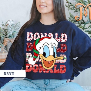 May include: Navy blue sweatshirt featuring a graphic of Donald Duck wearing a Santa hat. The word "DONALD" is repeated three times in a festive font with a Christmas pattern. The word "NAVY" is printed at the bottom.