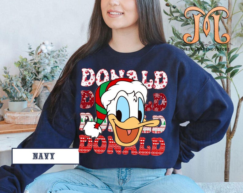 May include: Navy blue sweatshirt featuring a graphic of Donald Duck wearing a Santa hat. The word "DONALD" is repeated three times in a festive font with a Christmas pattern. The word "NAVY" is printed at the bottom.