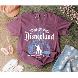 Puede incluir: Una camiseta granate con el texto "Walt Disney's Disneyland" en blanco y azul. El gr&aacute;fico incluye una silueta de castillo y la frase "The Happiest Place On Earth". La camiseta se muestra con zapatillas blancas, un sombrero y una manta azul.