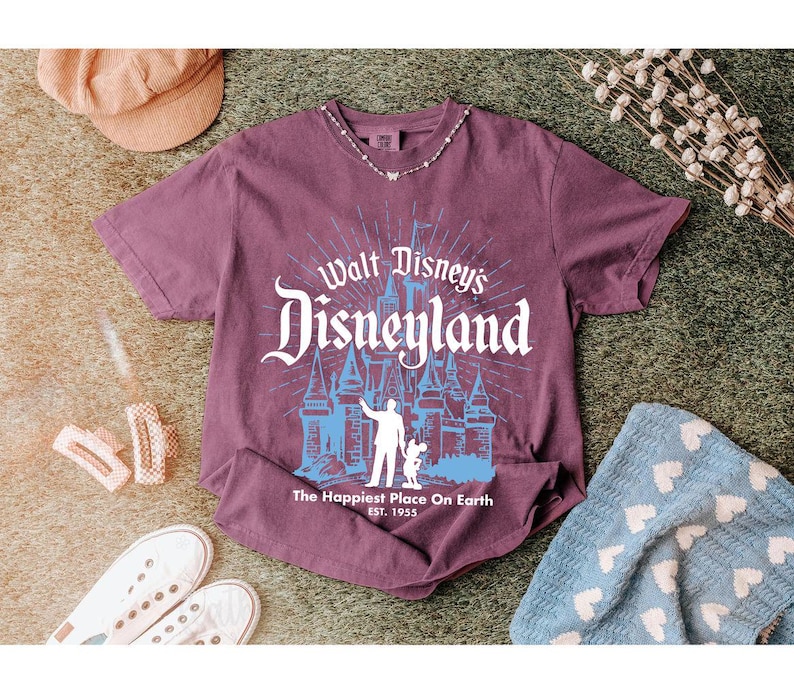 Puede incluir: Una camiseta granate con el texto "Walt Disney's Disneyland" en blanco y azul. El gr&aacute;fico incluye una silueta de castillo y la frase "The Happiest Place On Earth". La camiseta se muestra con zapatillas blancas, un sombrero y una manta azul.