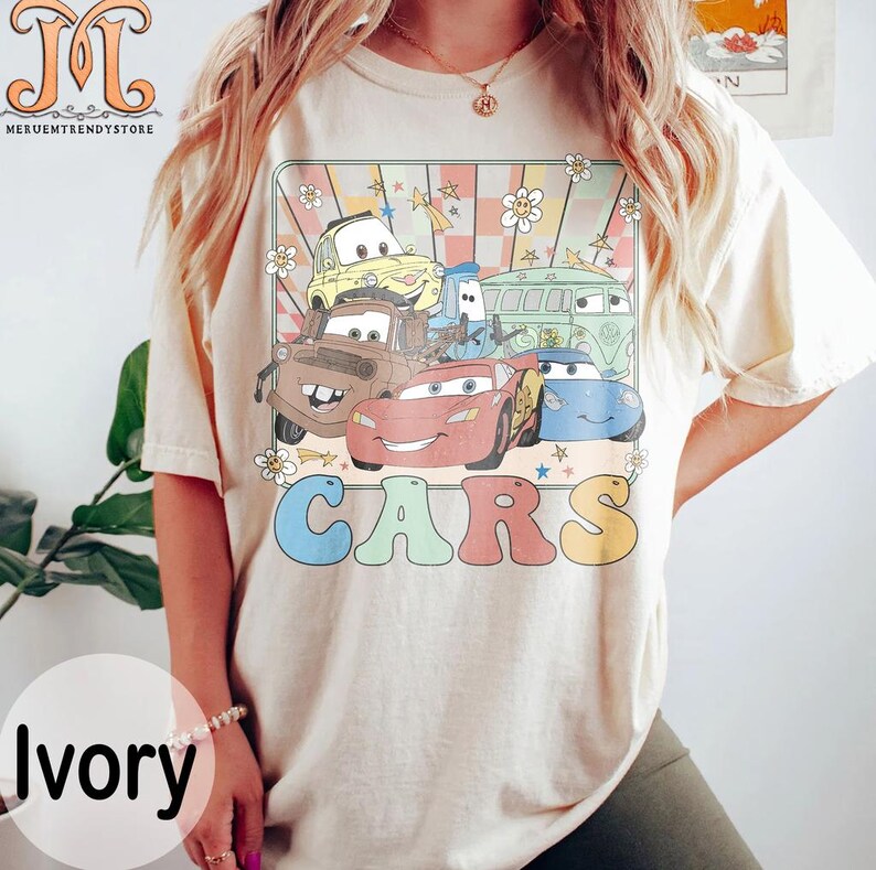 May include: A light beige t-shirt with a retro-style graphic of the Cars characters. The graphic features Lightning McQueen, Mater, and other Cars characters in a colorful, cartoon style. The word "CARS" is written in large, colorful letters below the graphic.