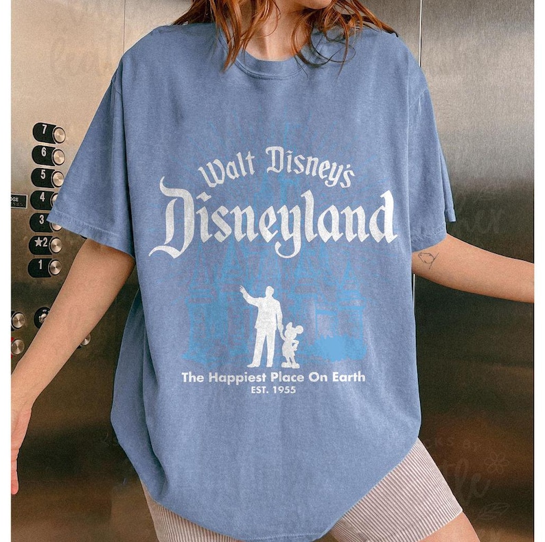 Puede incluir: Camiseta azul claro con la inscripci&oacute;n blanca "Walt Disney's Disneyland", una ilustraci&oacute;n de una persona y Mickey Mouse. La camiseta tambi&eacute;n incluye la frase "The Happiest Place On Earth" y "EST. 1955".