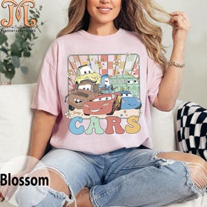 May include: A pink t-shirt with a retro-style graphic of the Cars characters. The graphic features Lightning McQueen, Mater, and other Cars characters in a colorful, checkered background with flowers and stars. The word "CARS" is written in large, colorful letters below the graphic.