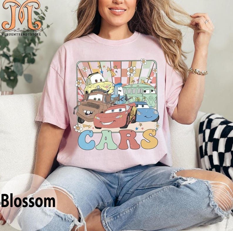 May include: A pink t-shirt with a retro-style graphic of the Cars characters. The graphic features Lightning McQueen, Mater, and other Cars characters in a colorful, checkered background with flowers and stars. The word "CARS" is written in large, colorful letters below the graphic.