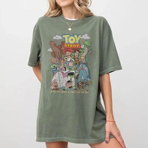 Puede incluir: Una camiseta verde oliva con un gr&aacute;fico colorido de personajes de la pel&iacute;cula animada "Toy Story". El dise&ntilde;o incluye el t&iacute;tulo de la pel&iacute;cula y la frase "You've Got a Friend in Me".