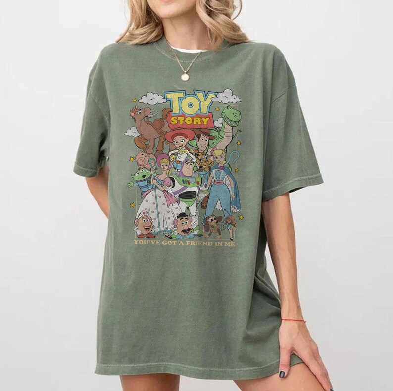 Puede incluir: Una camiseta verde oliva con un gr&aacute;fico colorido de personajes de la pel&iacute;cula animada "Toy Story". El dise&ntilde;o incluye el t&iacute;tulo de la pel&iacute;cula y la frase "You've Got a Friend in Me".