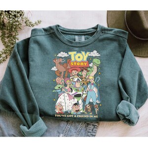 Puede incluir: Sudadera de color verde azulado con un estampado colorido de personajes de la pel&iacute;cula "Toy Story". El dise&ntilde;o incluye las palabras "Toy Story" y "You've Got a Friend in Me". La sudadera tiene mangas largas y cuello redondo.