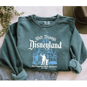 Puede incluir: Sudadera de cuello redondo color verde azulado con un dise&ntilde;o blanco y azul de Disneyland. El dise&ntilde;o incluye el castillo, una persona y Mickey Mouse. El texto dice "Walt Disney's Disneyland" y "The Happiest Place On Earth EST. 1955".