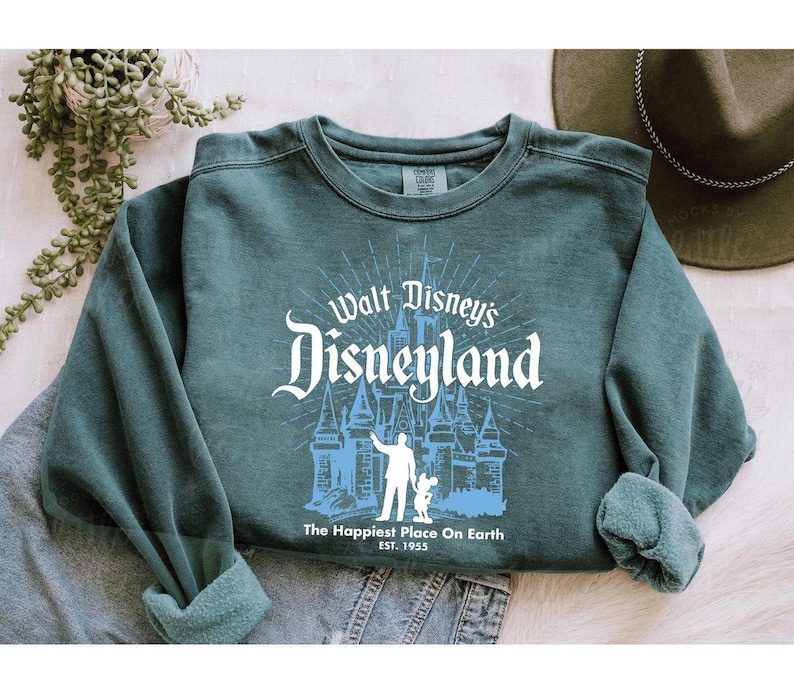 Puede incluir: Sudadera de cuello redondo color verde azulado con un dise&ntilde;o blanco y azul de Disneyland. El dise&ntilde;o incluye el castillo, una persona y Mickey Mouse. El texto dice "Walt Disney's Disneyland" y "The Happiest Place On Earth EST. 1955".