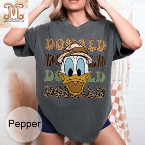 May include: A dark gray t-shirt with a graphic print of Donald Duck wearing a safari hat. The graphic is surrounded by a leopard print pattern and the text "DONALD" is repeated three times.