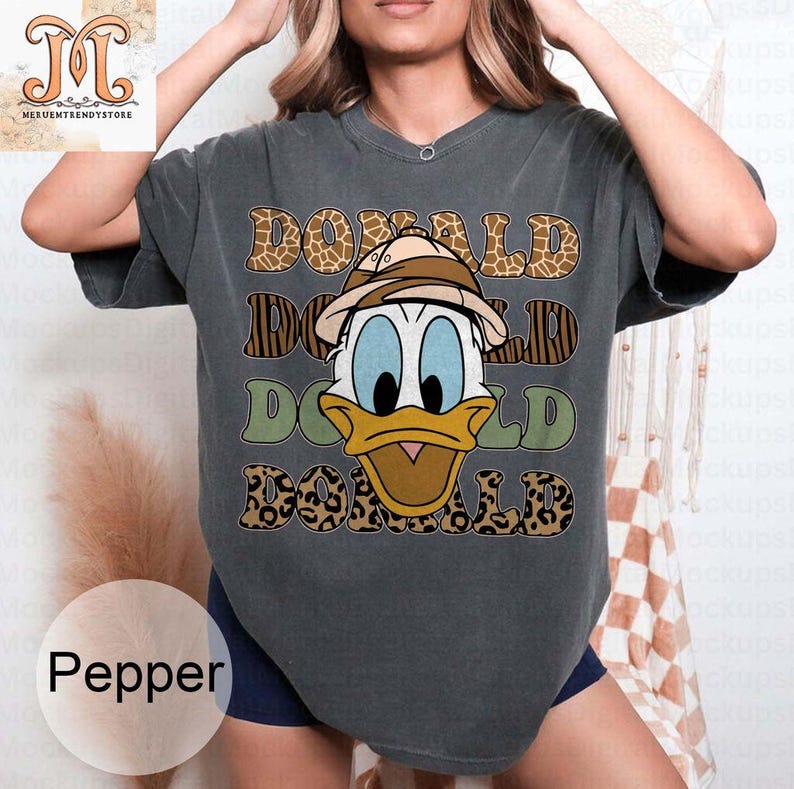 May include: A dark gray t-shirt with a graphic print of Donald Duck wearing a safari hat. The graphic is surrounded by a leopard print pattern and the text "DONALD" is repeated three times.