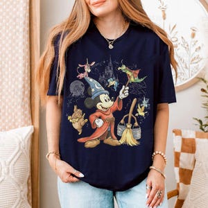 K&ouml;nnte beinhalten: Marineblaues T-Shirt mit Mickey Mouse als Zauberer, der einen Zauberstab h&auml;lt und von animierten Figuren und einem Schloss umgeben ist. Das Design beinhaltet einen Besen und einen Eimer, mit einer farbenfrohen Illustration.